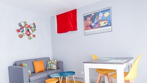 A modern living room with a grey sofa, colorful cushions, a red curtain, a framed abstract painting, a small table with chairs, and a yellow rug.