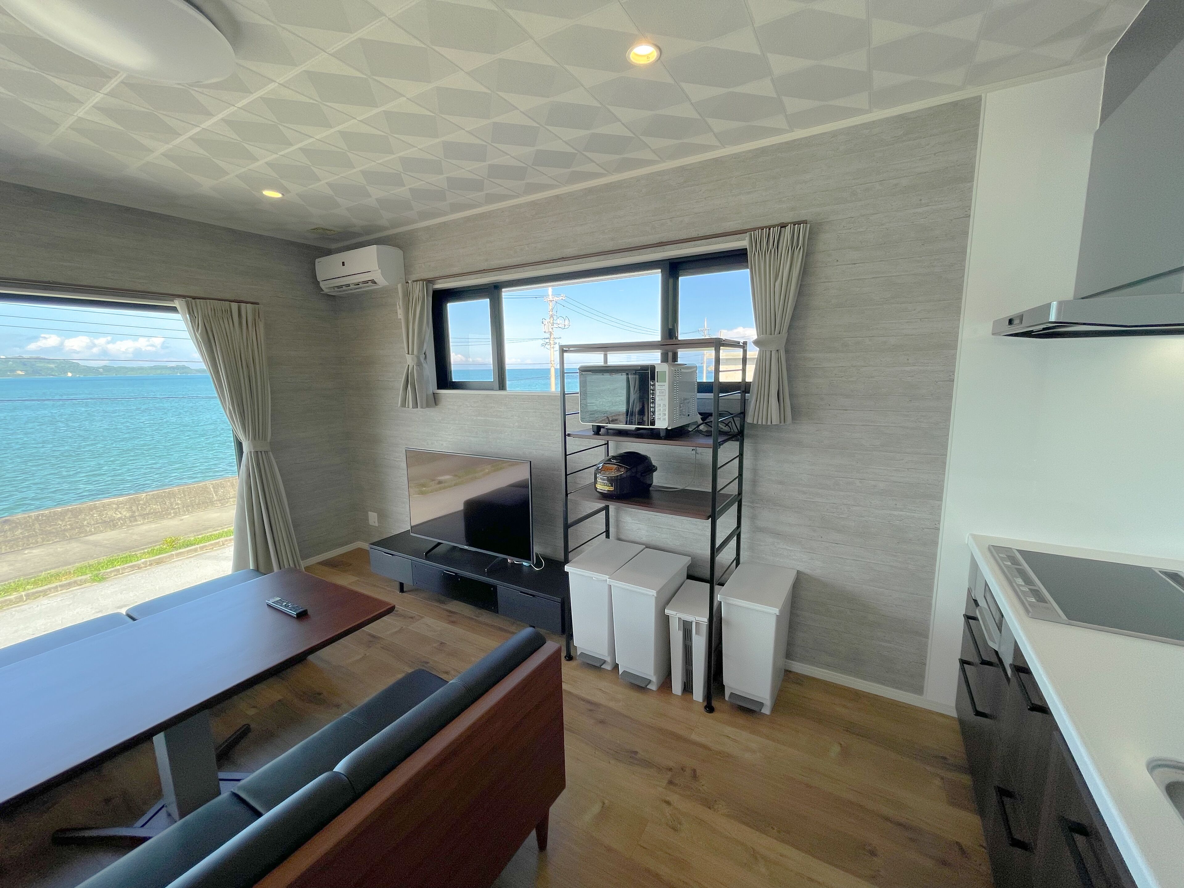 A compact room with a wooden floor, a dining table, a sofa, a kitchenette with a microwave, and a window overlooking the sea.