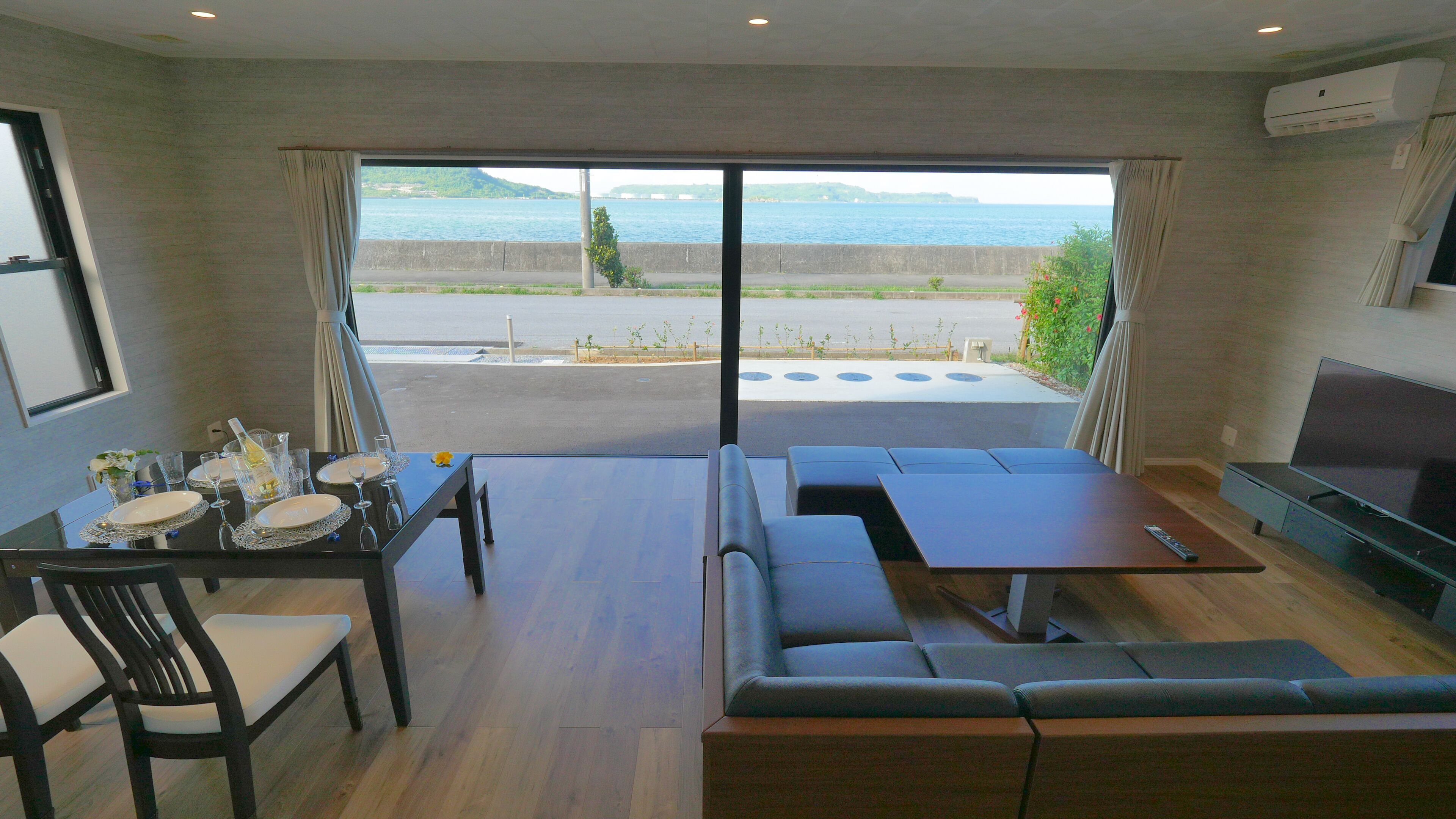 Una habitación con una mesa de comedor preparada para cuatro personas, un sofá, un televisor y vista al mar.