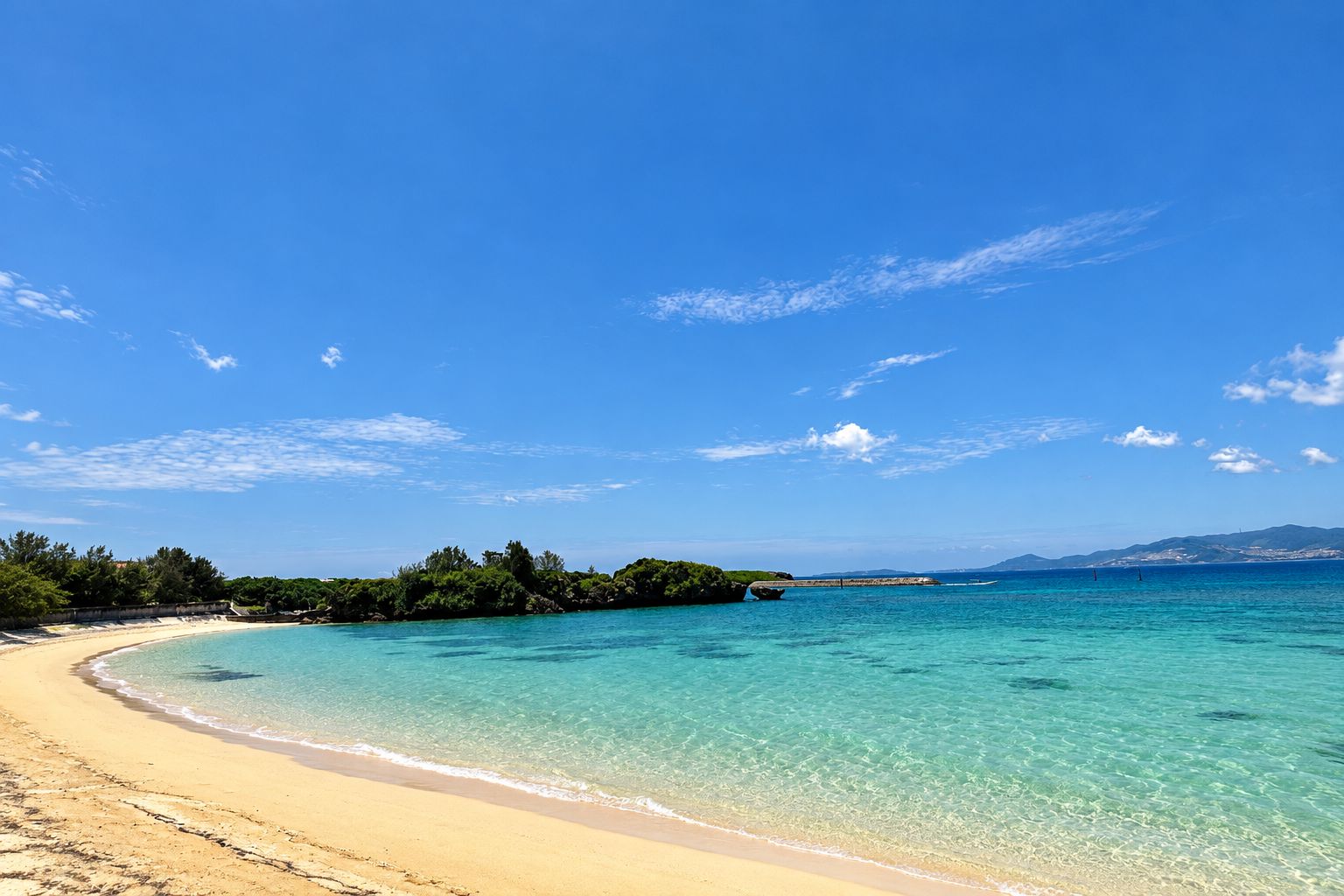 A beach with clear turquoise water, sandy shore, and a distant bridge.