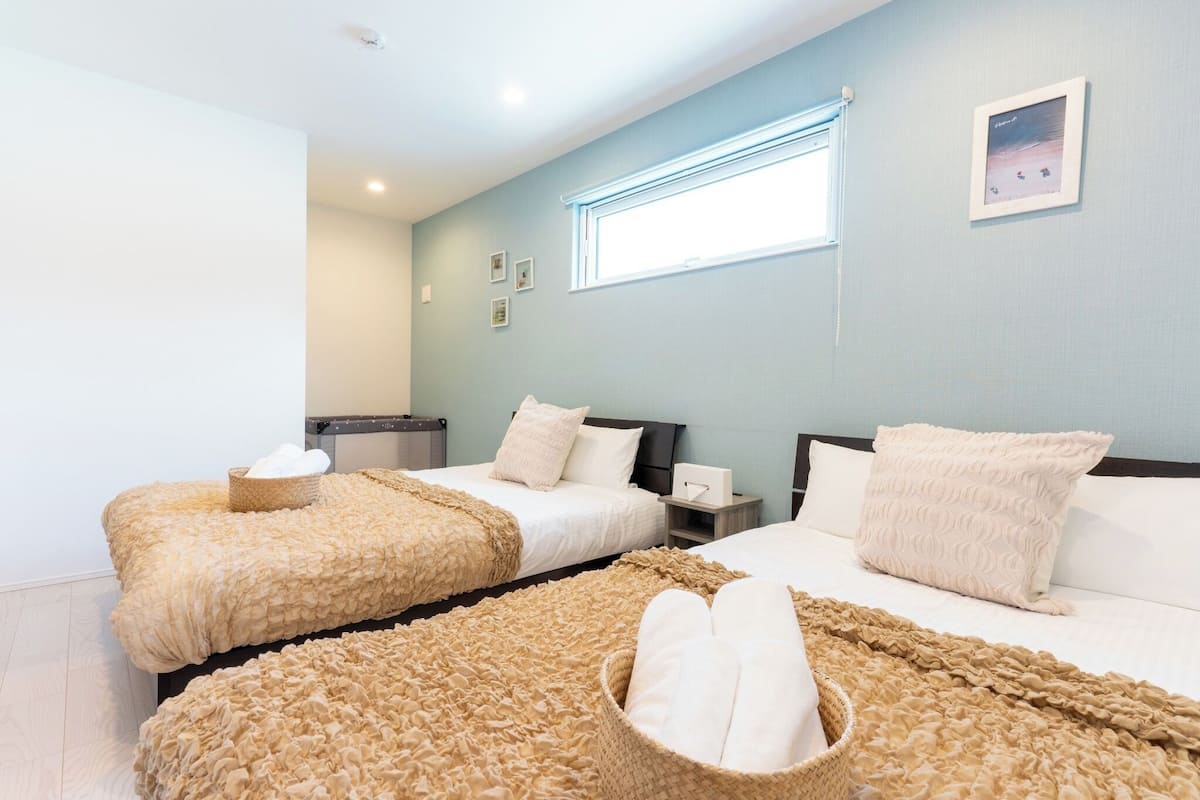 A hotel room with two beds, each with a beige textured blanket and pillows, a window with blinds, and a framed picture on the wall.