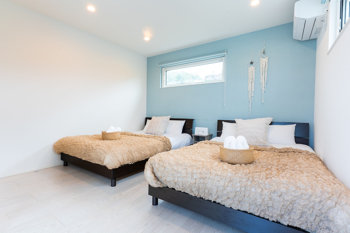 Une chambre avec deux lits, chacun recouvert d’une couverture à volants et agrémenté d’un panier. Il y a une fenêtre donnant sur les arbres, et un climatiseur fixé au mur.