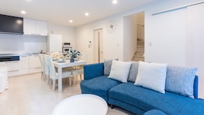 A modern living room with a blue sofa, white dining table, and kitchen area.