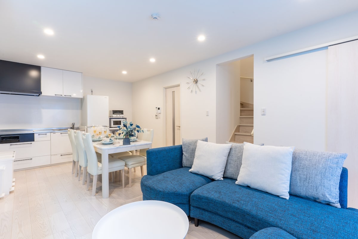 A modern living room with a blue sofa, white dining table, and kitchen area.