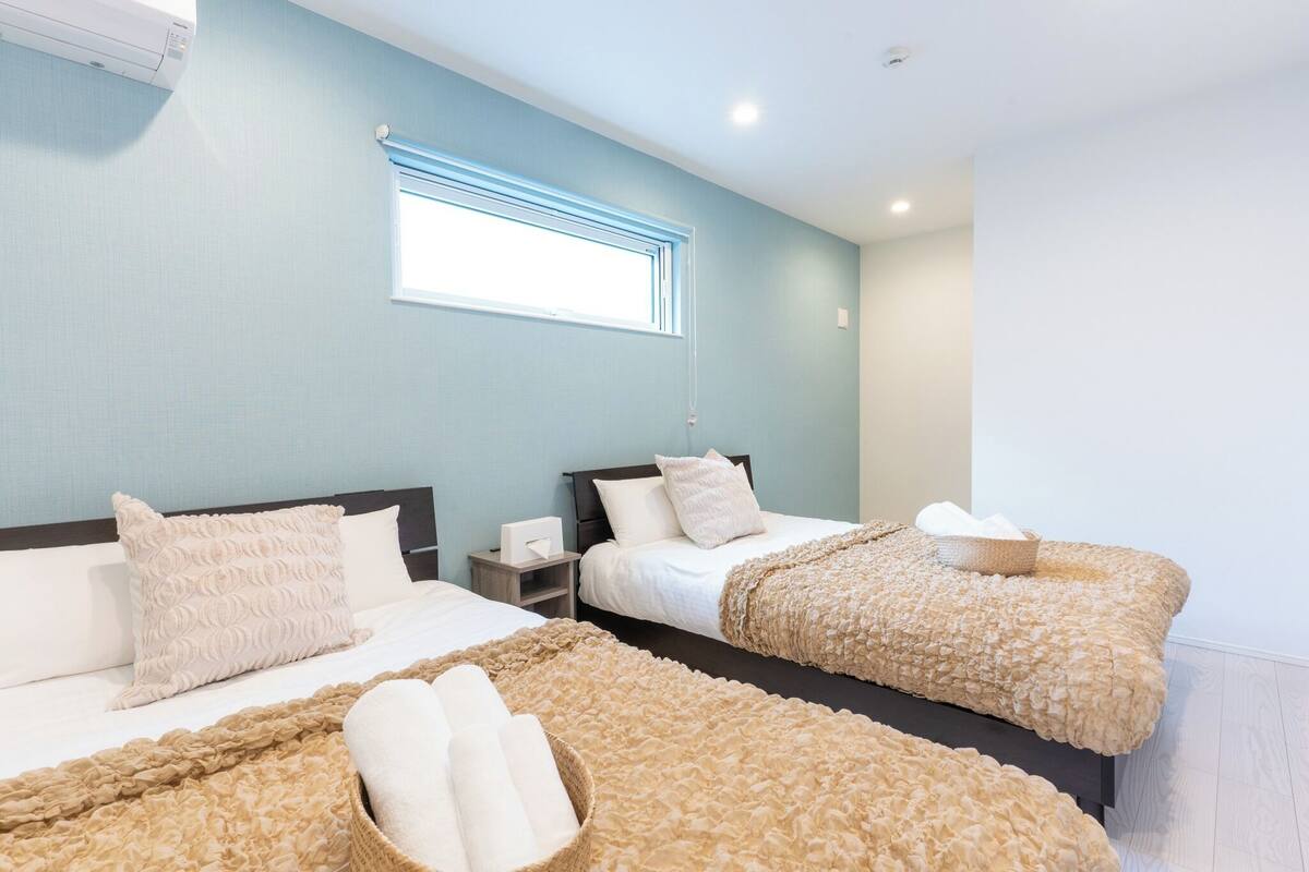 A hotel room with two beds, a window with blinds, a bedside table with a lamp, and a wall-mounted air conditioner.