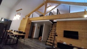Un intérieur moderne avec une structure de plafond en bois, un coin repas comprenant des chaises et une table, un escalier et un téléviseur à écran plat fixé au mur.