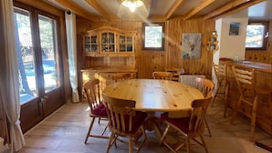 Interior de madera con una mesa redonda rodeada de sillas, una zona de cocina con armarios de madera, y un amplio ventanal que proporciona luz natural.