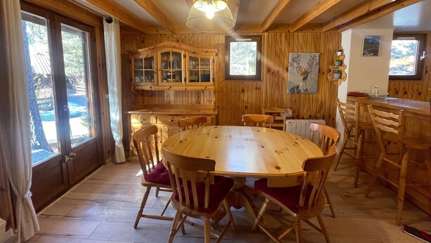 Interior de madera con una mesa redonda rodeada de sillas, una zona de cocina con armarios de madera, y un amplio ventanal que proporciona luz natural.