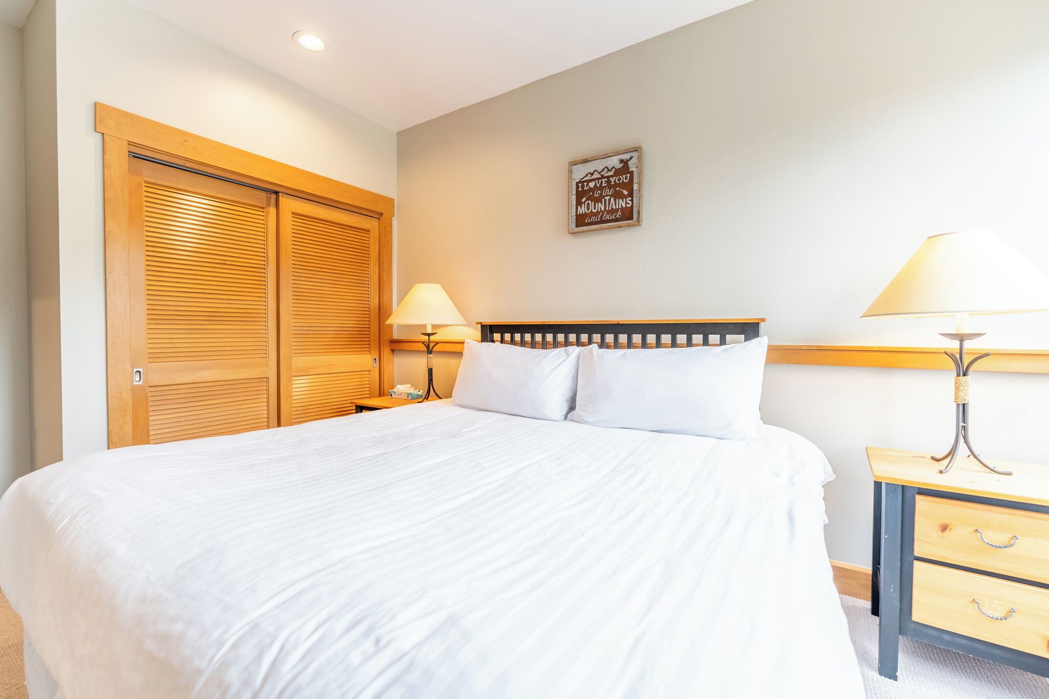 A bedroom with a large bed, wooden wardrobe, bedside tables with lamps, and a wall-mounted sign.