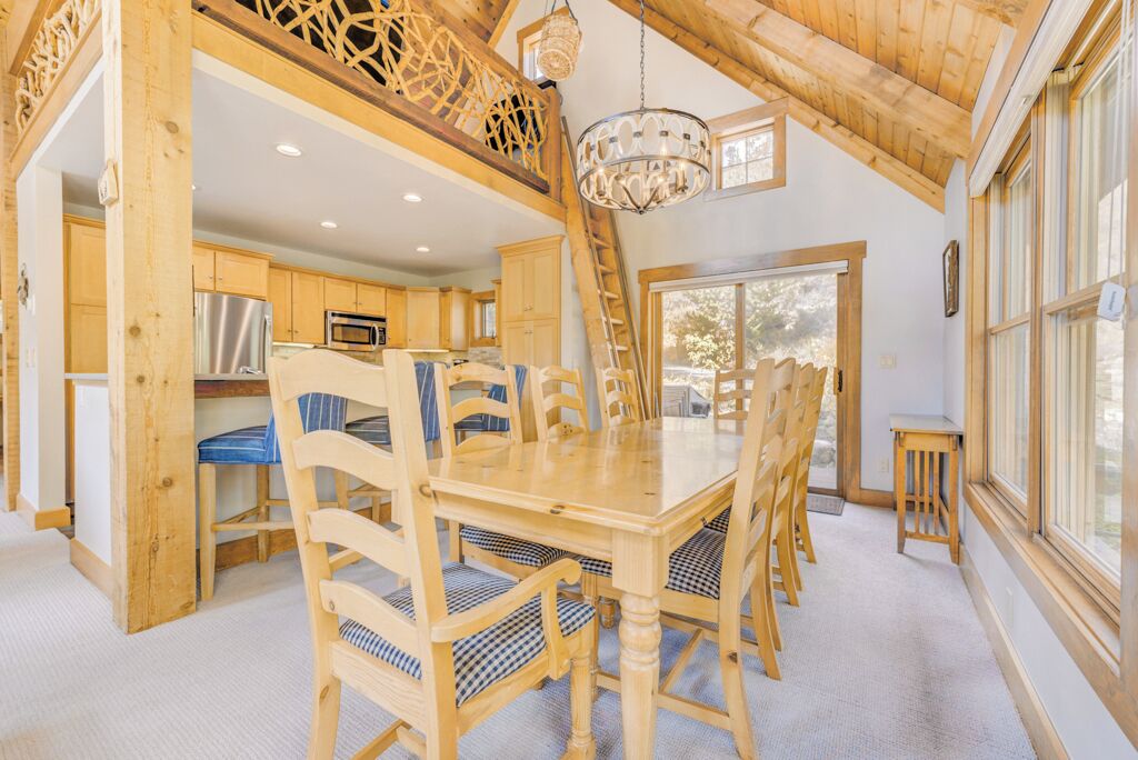 Una mesa de comedor de madera con sillas, una araña y vistas al exterior a través de amplios ventanales.
