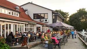 An outdoor dining area with tables and chairs, a building with a tiled roof, and a walkway along a water body. - Vacation with dog, and without dog (Varel)