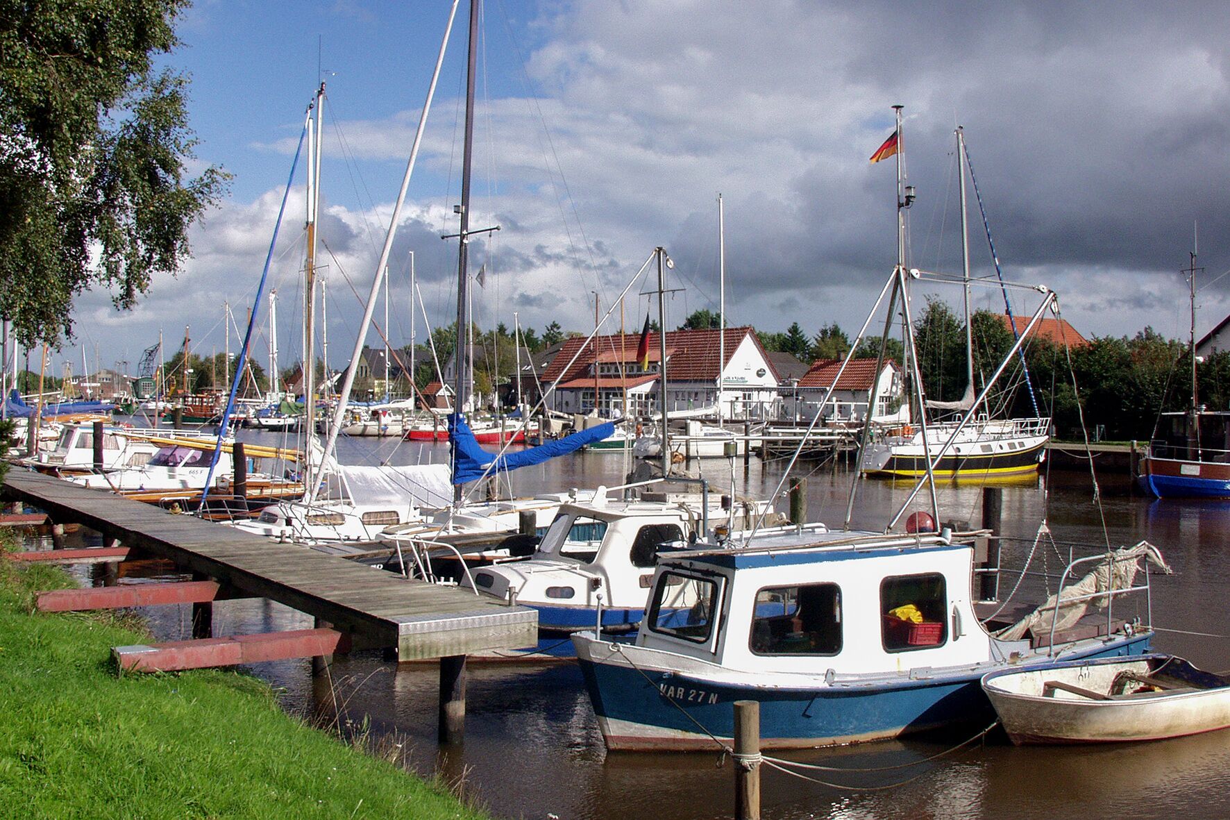 A marina with several boats docked, a wooden pier, and houses in the background.