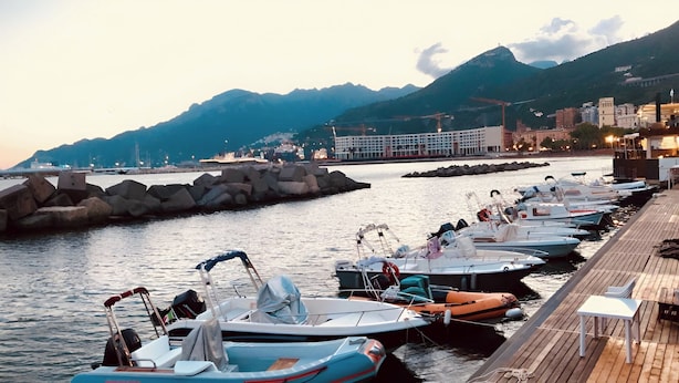 A marina with boats docked along a wooden pier, a rocky breakwater, and a cityscape with buildings and mountains in the background. - Arechi Room (Salerno)