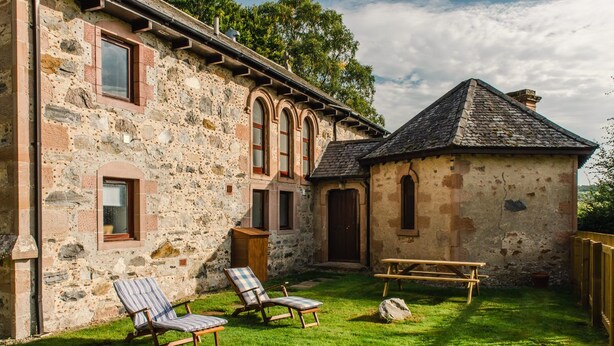 A stone building with a small attached structure, a grassy yard, and two lounge chairs.