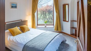 A bedroom with a wooden ceiling, a bed with white bedding, a window with curtains, and a view of trees.