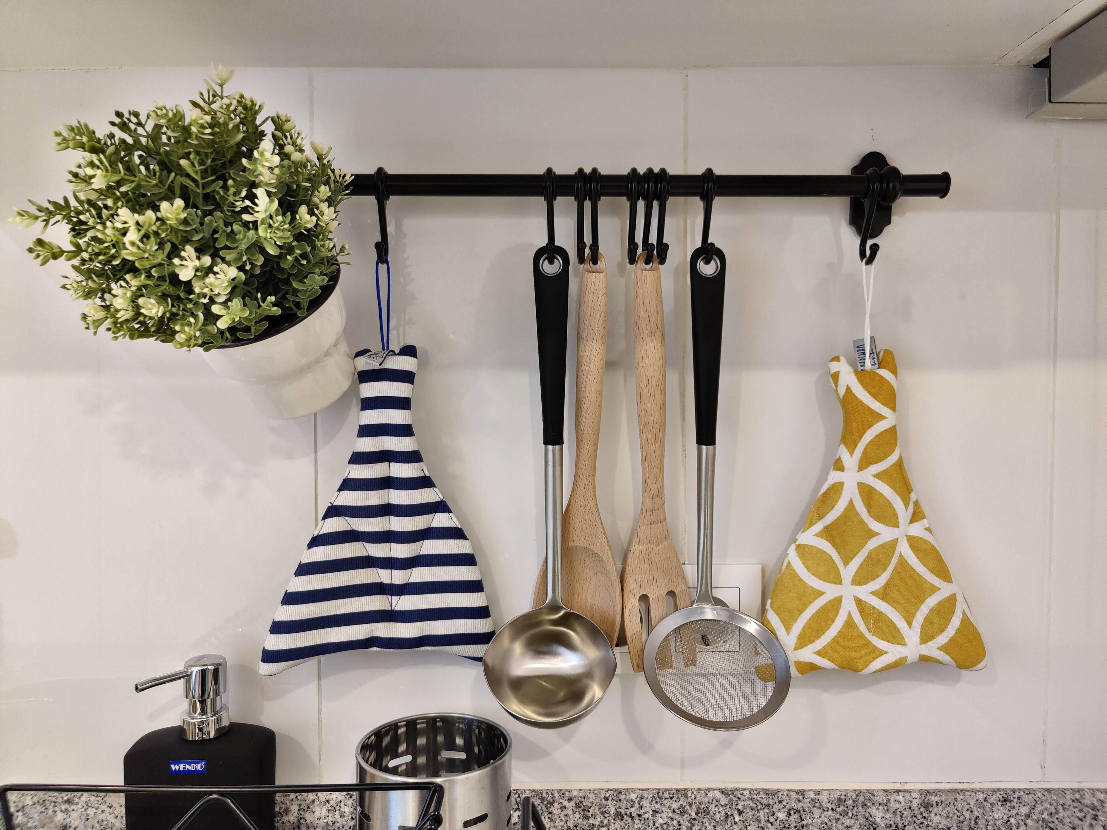 Una cocina con una planta colgante, un delantal a rayas, cucharones de madera y un agarradera amarilla estampada, además de una olla de acero inoxidable.