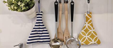 Una cocina con una planta colgante, un delantal a rayas, cucharones de madera y un agarradera amarilla estampada, además de una olla de acero inoxidable.