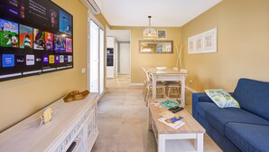 A living room with a TV displaying various streaming services, a wooden coffee table with magazines, a blue sofa, and a dining area with a table and chairs. - An oasis in the center of Corralejo! (Corralejo - Fuerteventura)