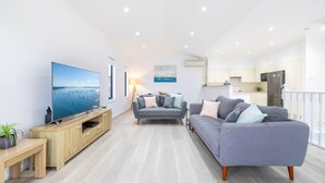 A modern living room with a large flat-screen TV, two grey sofas, a wooden coffee table, and a small side table with a plant. - Resthaven, 3b Messines Street (Shoal Bay)