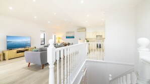 A spacious living room with a large flat-screen TV, a grey sofa, and a wooden coffee table. There is a staircase leading to an upper level with a kitchen area visible. - Resthaven, 3b Messines Street (Shoal Bay)