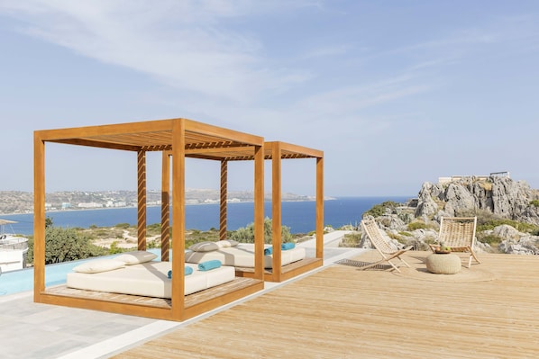 Une terrasse en bois agrémentée de deux pergolas en bois, une piscine et une vue sur la mer et le paysage rocheux.