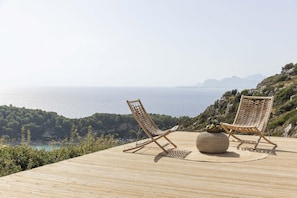 Une terrasse en bois agrémentée de deux fauteuils de jardin en osier et d’un pouf tressé, offrant une vue sur un paysage côtier avec des falaises et la mer.