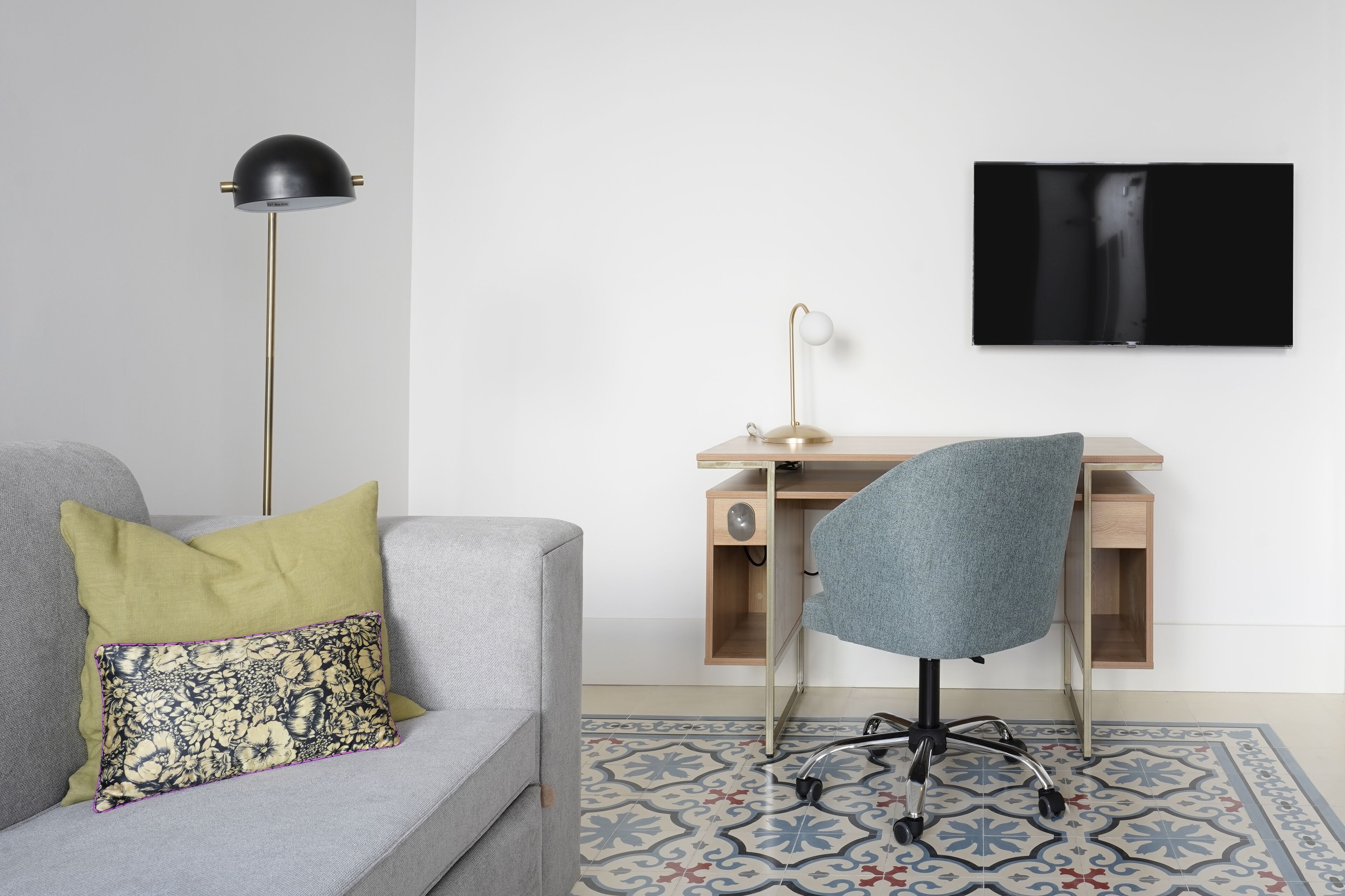 Un salon moderne avec un canapé gris, un coussin à motifs, une lampe sur pied, un bureau avec une chaise et un téléviseur à écran plat fixé au mur.