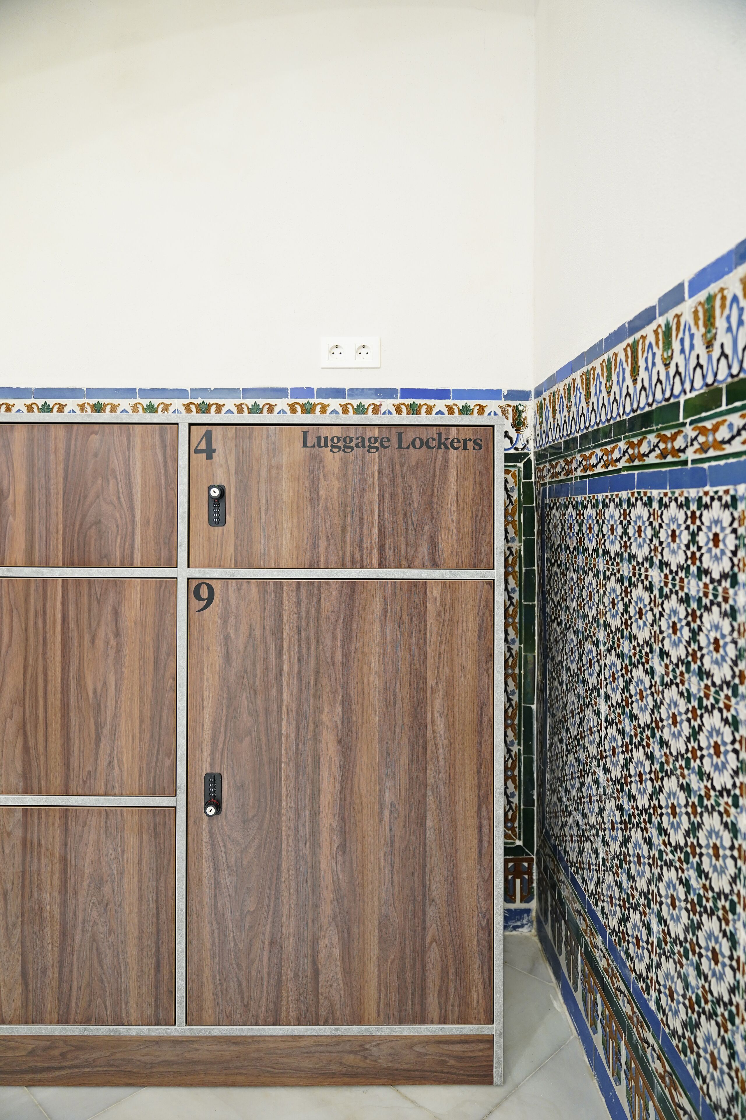 A set of wooden luggage lockers with numbers 4 and 9, a wall-mounted electrical outlet, and a decorative tiled wall.
