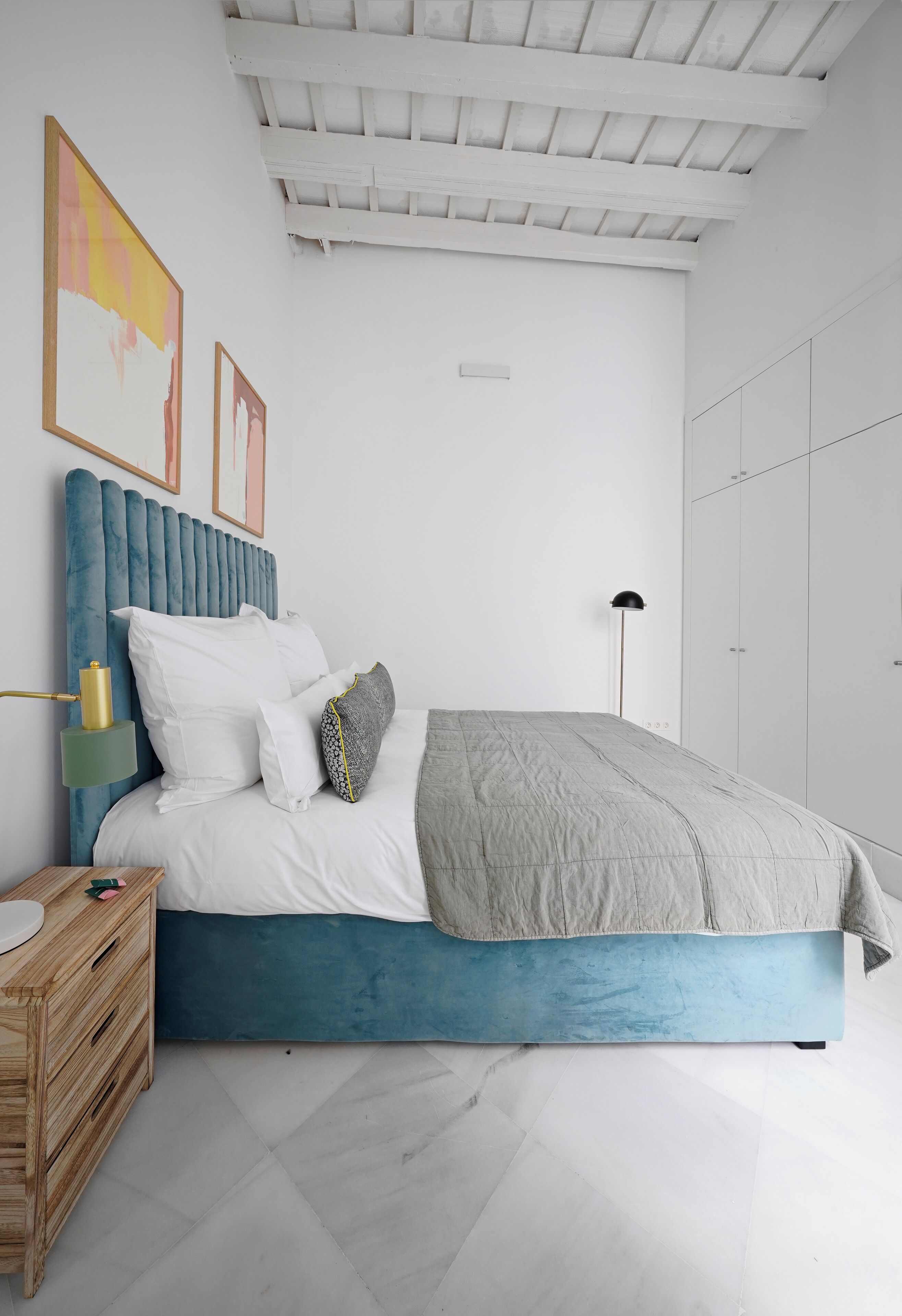 A bedroom with a blue upholstered headboard, white bedding, a bedside table with a lamp, and a wardrobe.