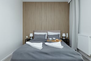 A neatly made bed with gray bedding, two pillows, and two folded towels. There is a tray with a cup and a pastry on the bed.