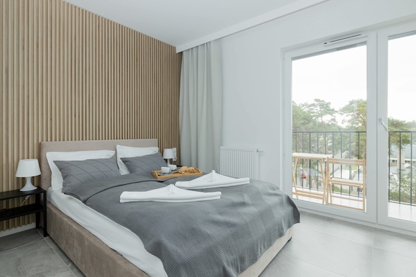 A cozy bedroom featuring a double bed with gray bedding and a wooden accent wall behind it. Large windows offer natural light and a view of greenery.