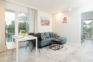 A modern living room with a sofa, a dining table set for two, and a view of the outdoors through large windows.