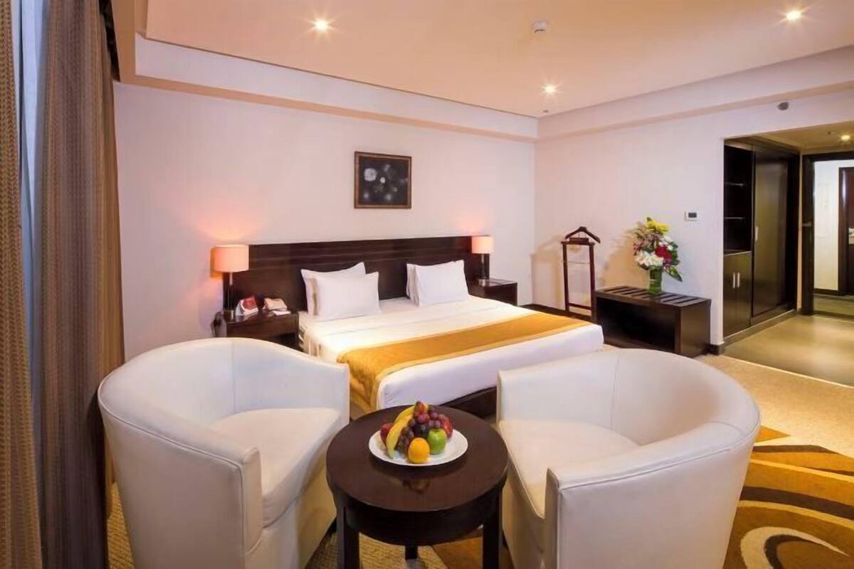 A hotel room with a bed, two white armchairs, a small table with fruit, a nightstand, and a lamp.