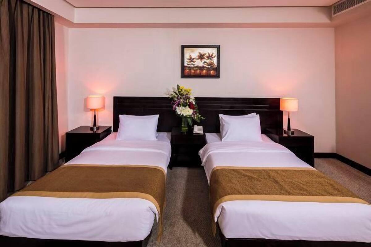 A hotel room with two beds, a nightstand with a lamp, a vase with flowers, and a framed picture on the wall.