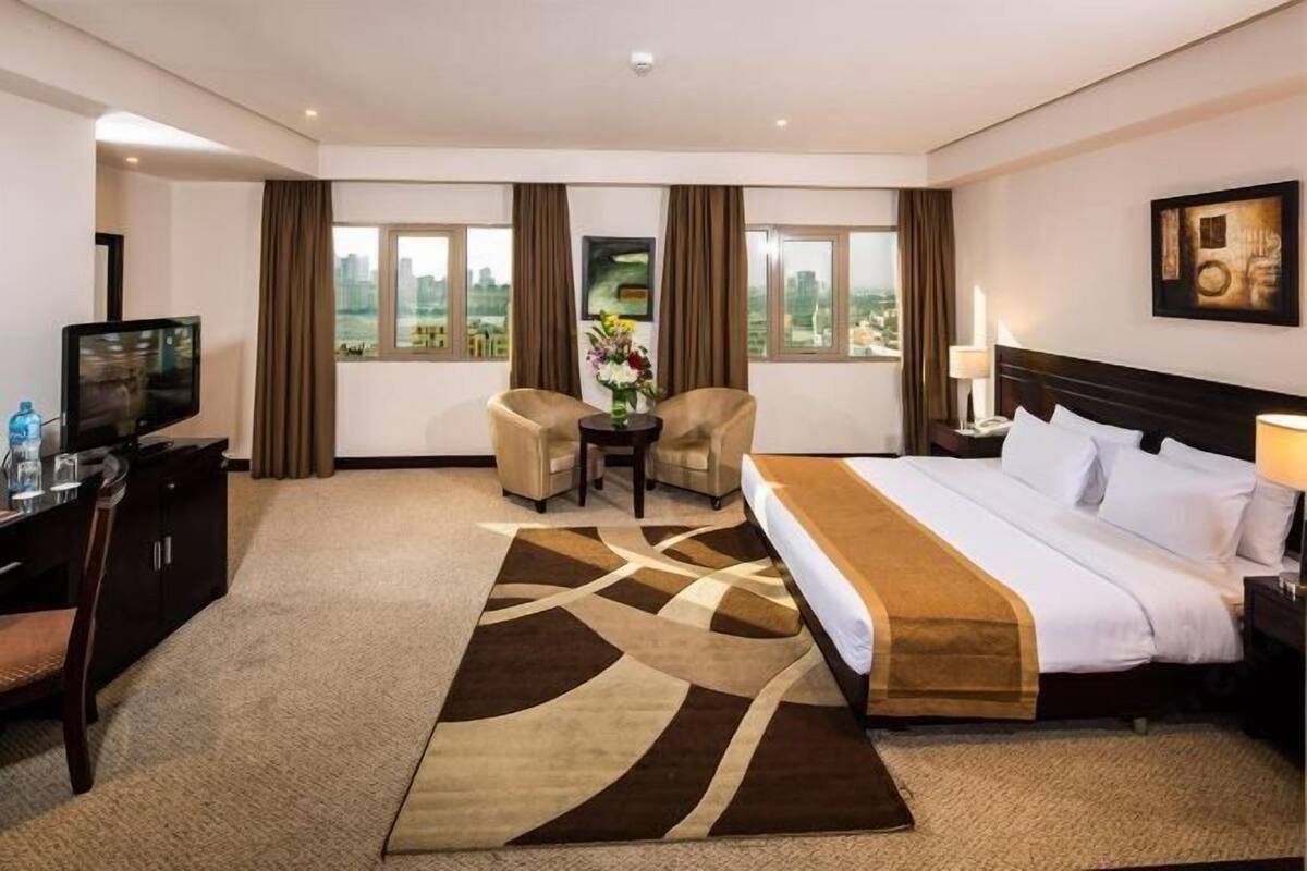 A hotel room with a bed, two armchairs, a small table with a vase, a TV, and a city view through large windows.
