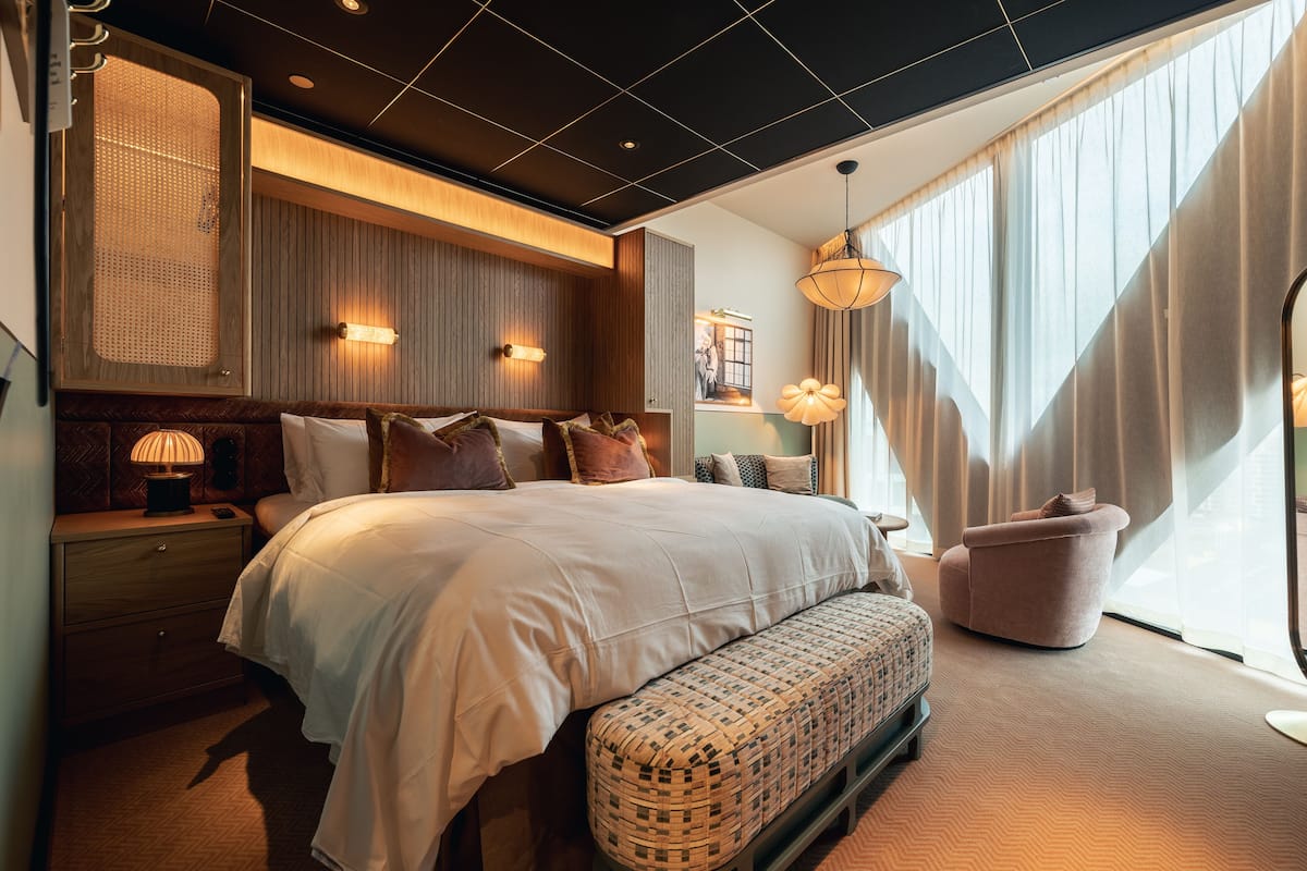 A modern hotel room with a large bed, a bench, a nightstand, and a window with curtains.