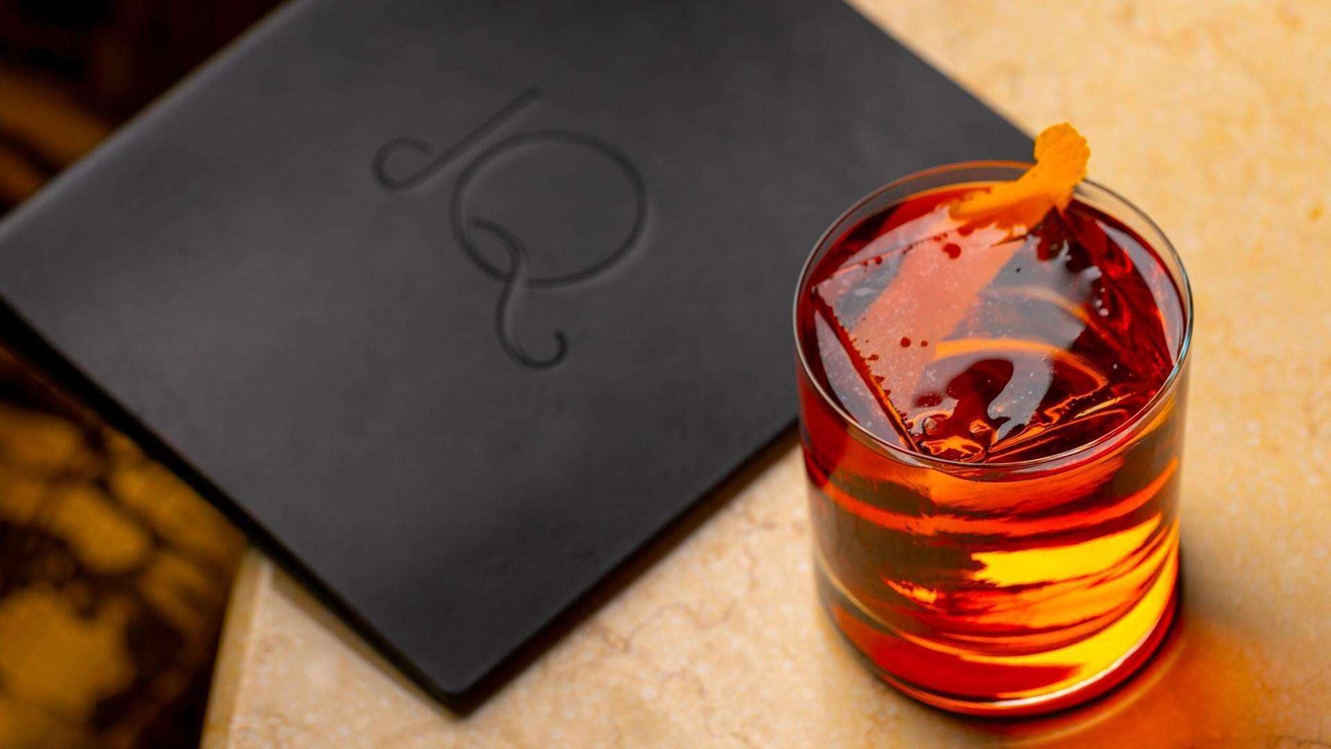 Ein Glas bernsteinfarbenes Getränk mit Garnitur, neben einer schwarzen Kiste mit einem Logo platziert.