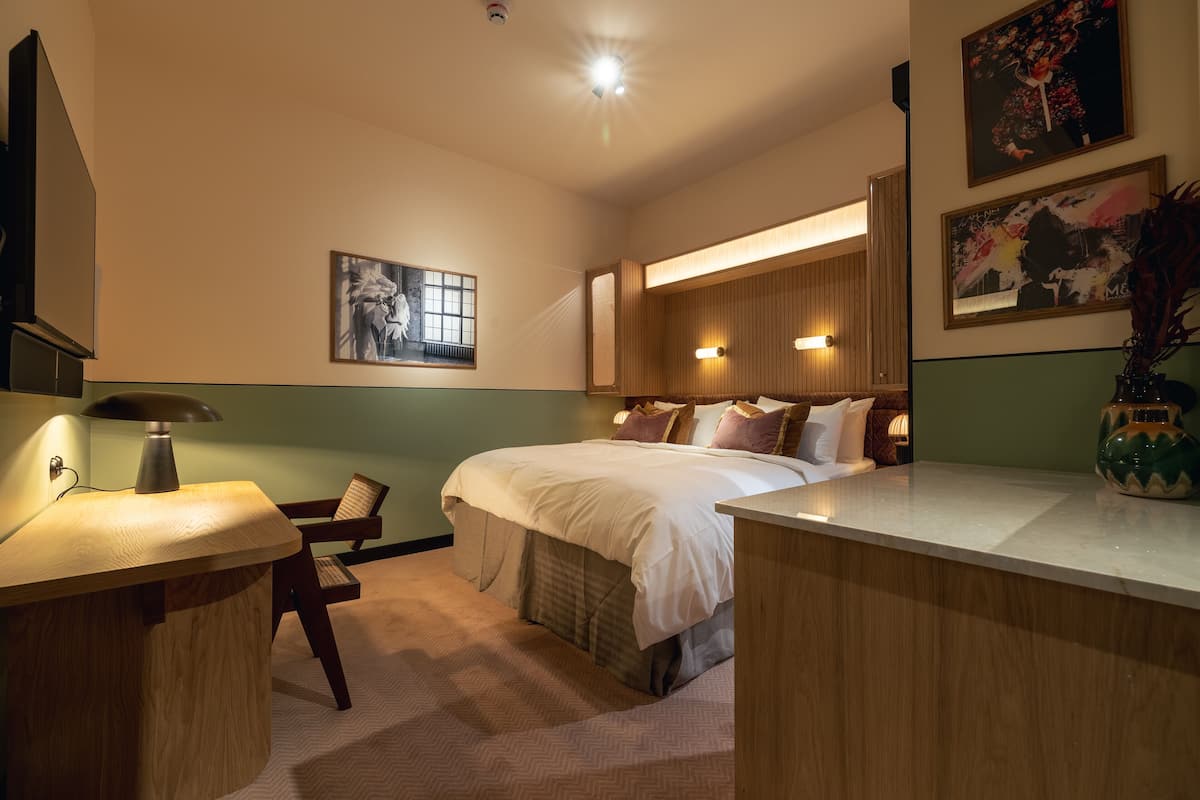 A hotel room with a bed, bedside table, lamp, chair, artwork, and a decorative vase.