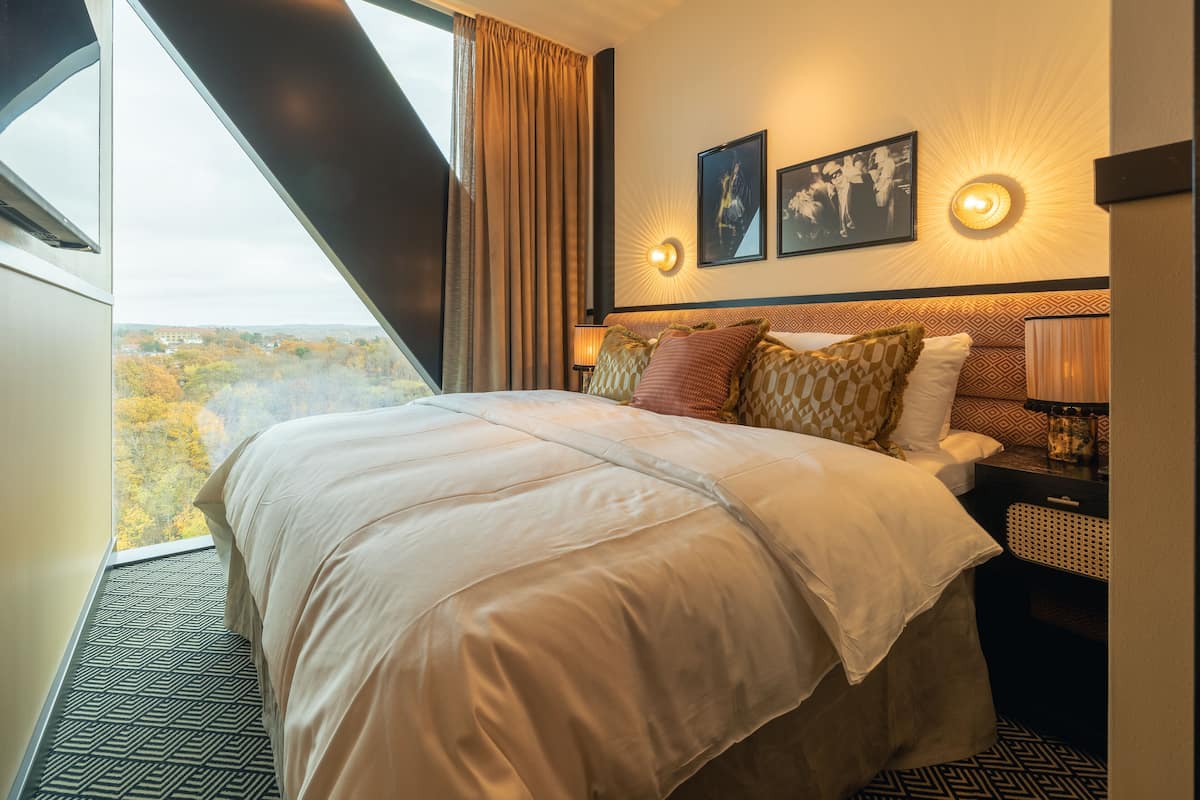 A hotel room with a large bed, a nightstand, a lamp, and a view of the outdoors.