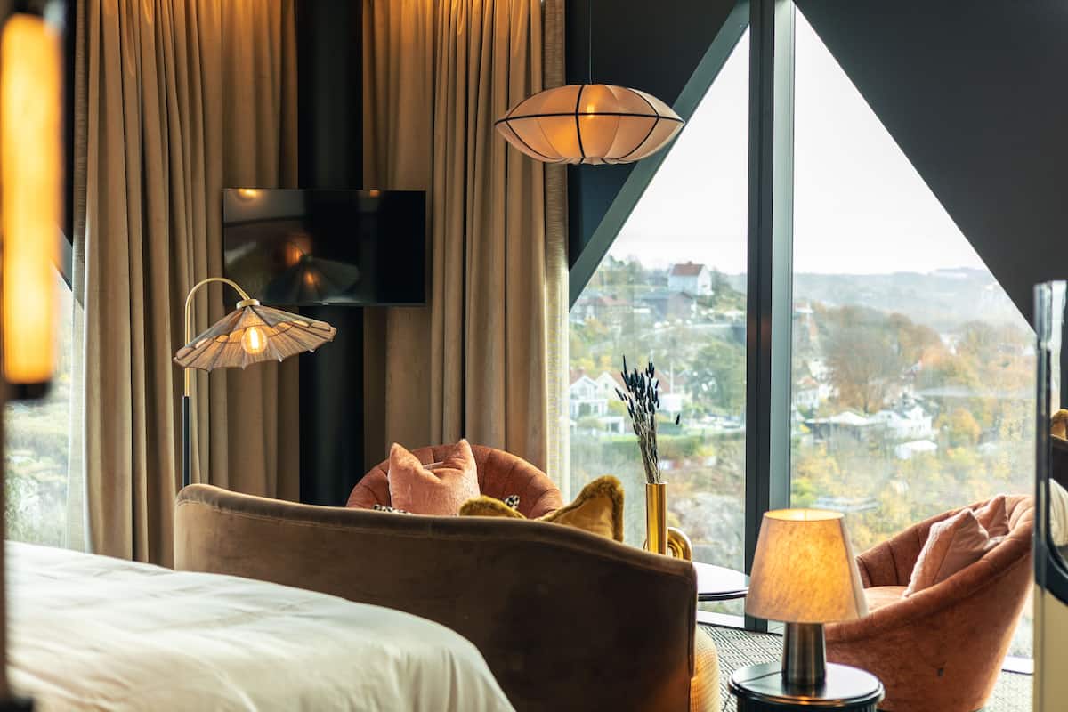 A hotel room with a large window, a bed, a chair, and a lamp.