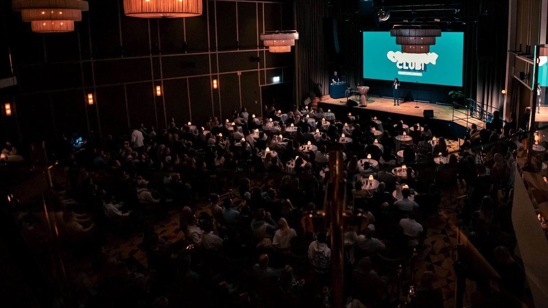 A large indoor gathering with a stage and a screen displaying 'CLUB' in the background.
