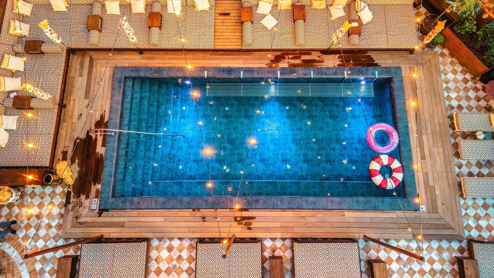 Una piscina rectangular con un deck de madera que la rodea, con un piso de patrón escocés y luces colgantes.
