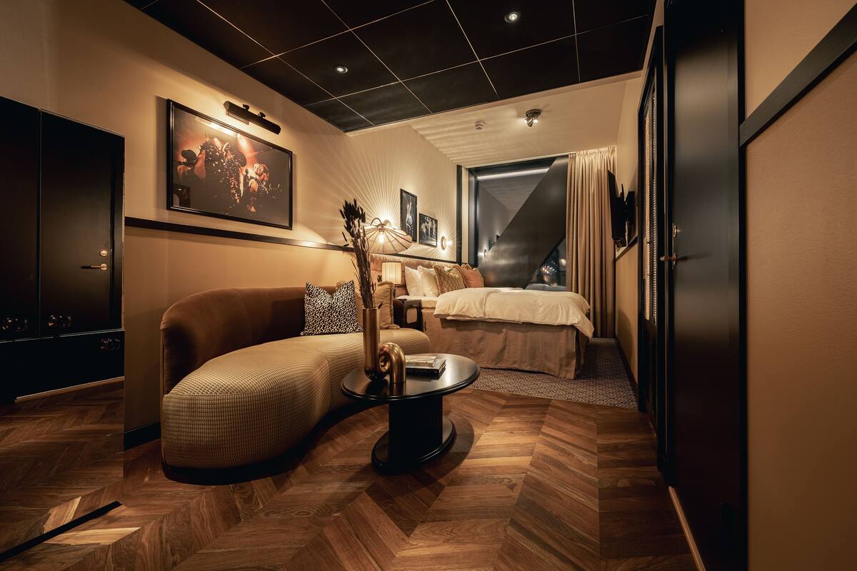 A modern hotel room with a sofa, a small round table, a bed, and a large window.