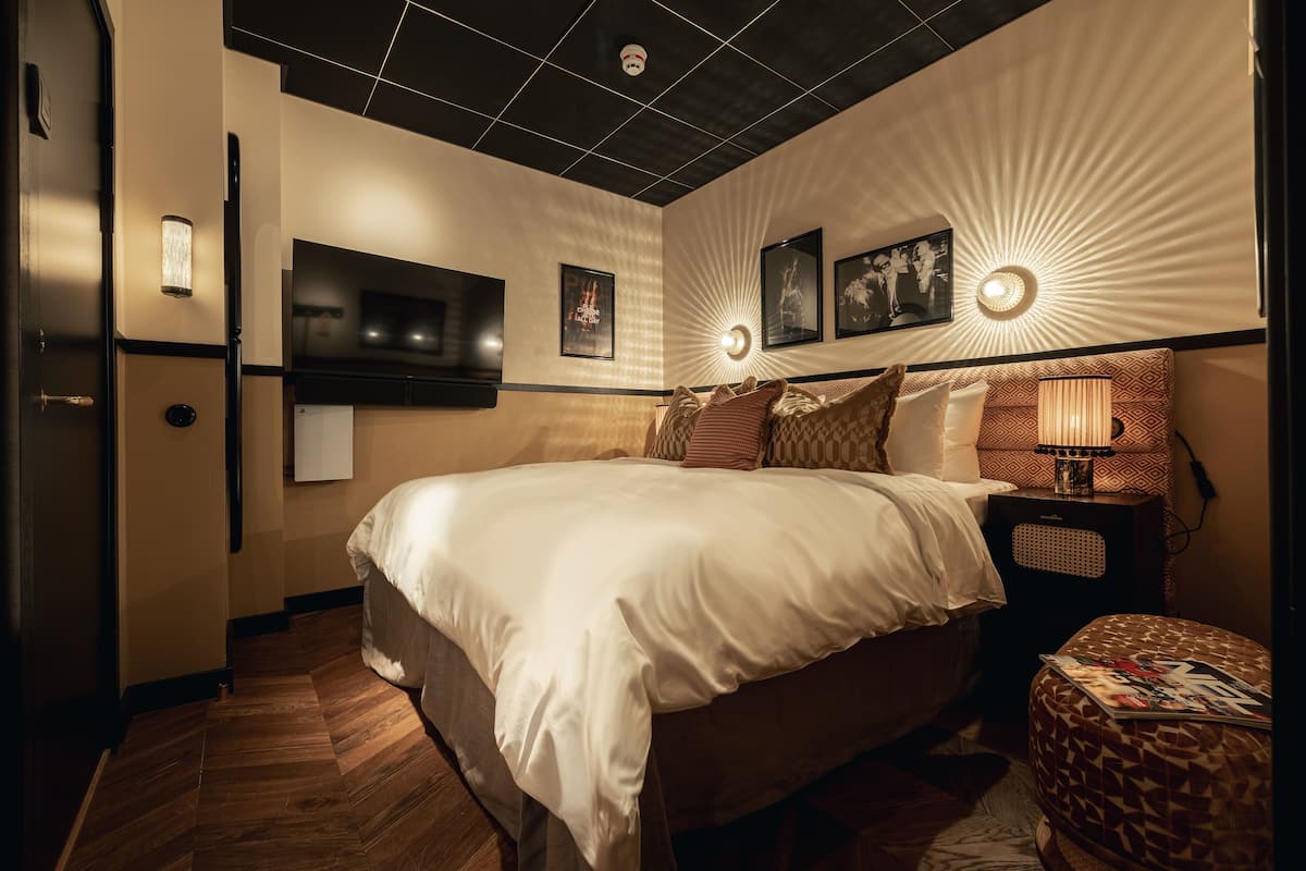 A hotel room with a large bed, a flat-screen TV mounted on the wall, a bedside table with a lamp, and a patterned ottoman.