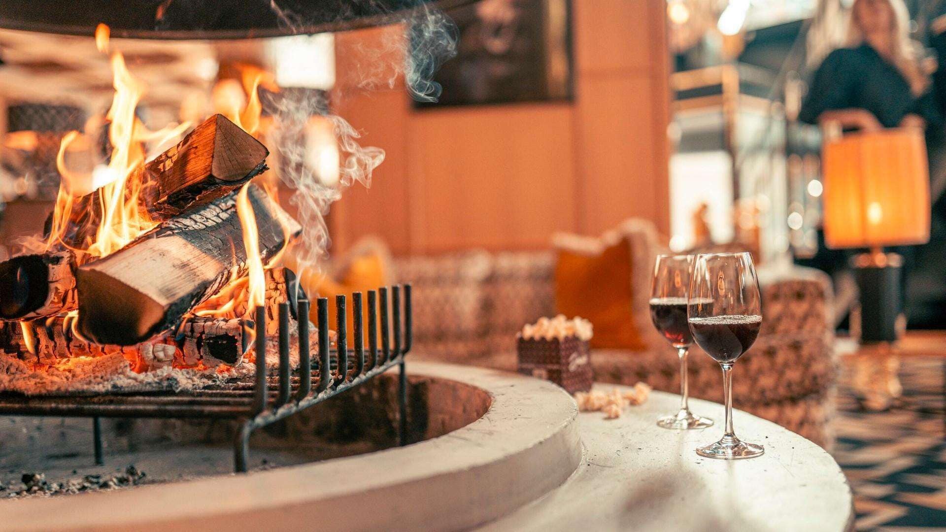 A fireplace with burning logs, two glasses of red wine, and a cozy seating area with cushions.