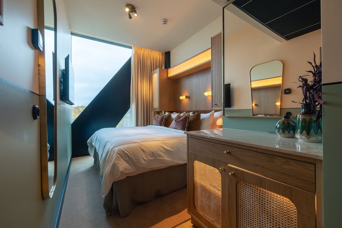 A modern hotel room with a bed, a wooden dresser, a mirror, and a view of the outdoors.