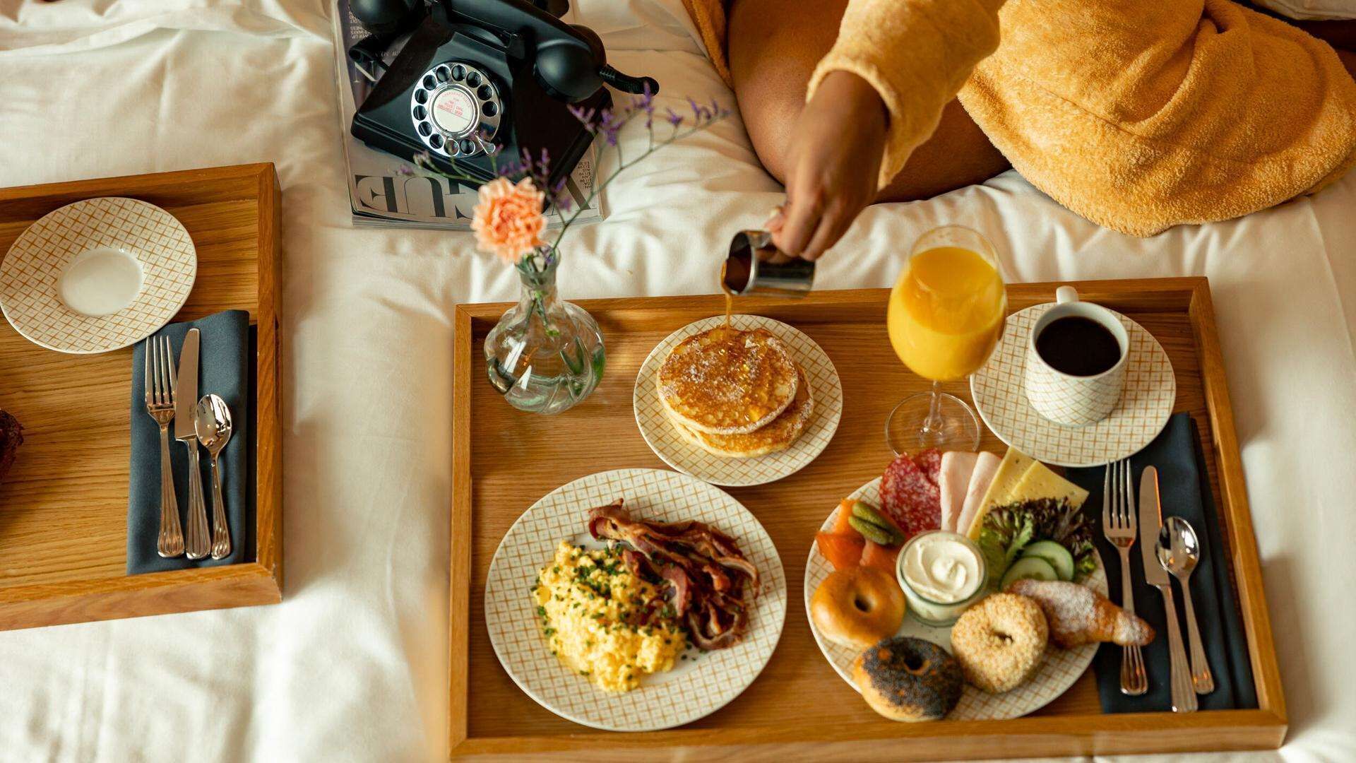 A breakfast tray with pancakes, eggs, bacon, bagels, and juice on a bed.