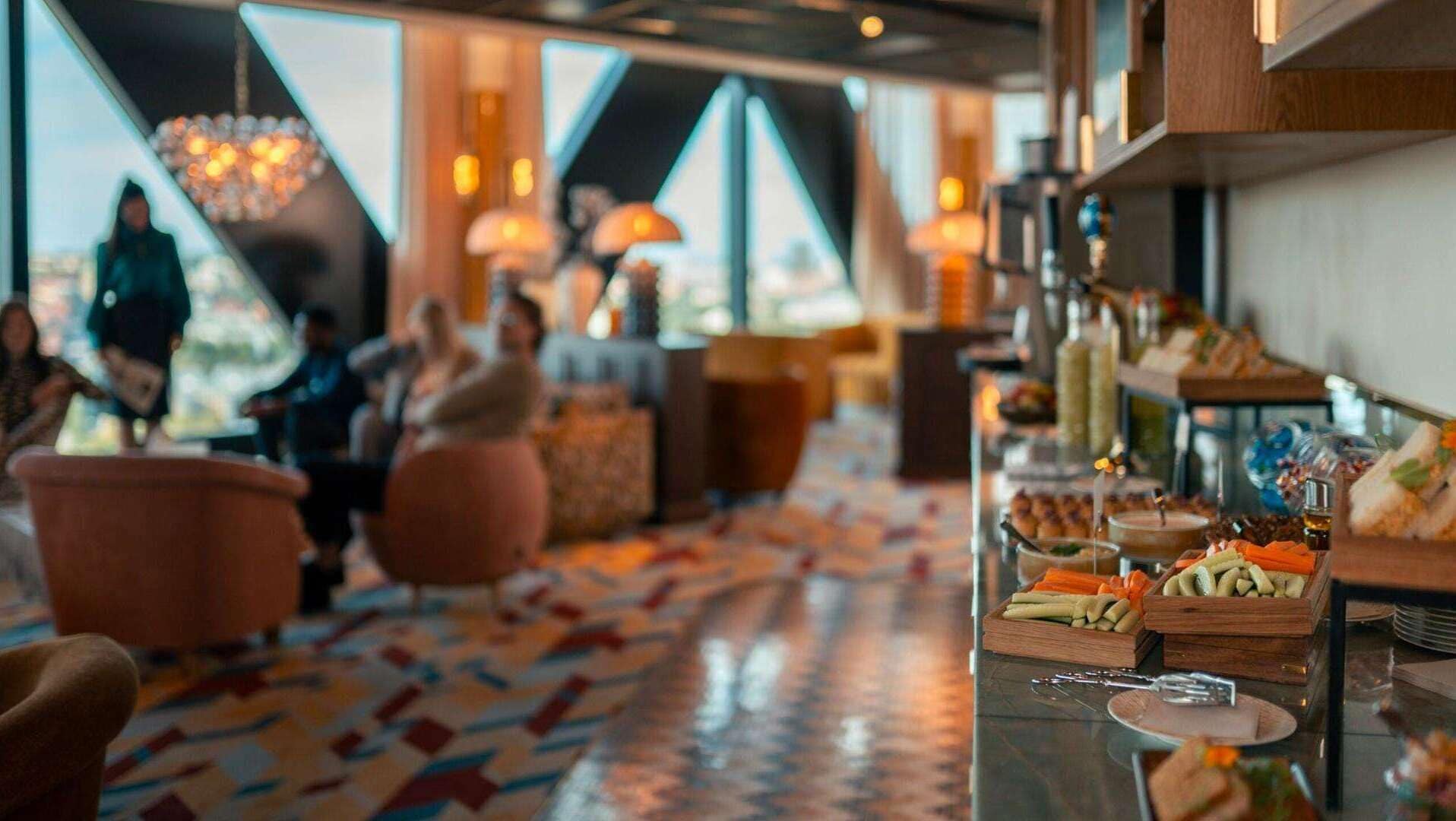 A hotel lobby with a buffet area featuring various food items and a seating area with chairs and a patterned floor.