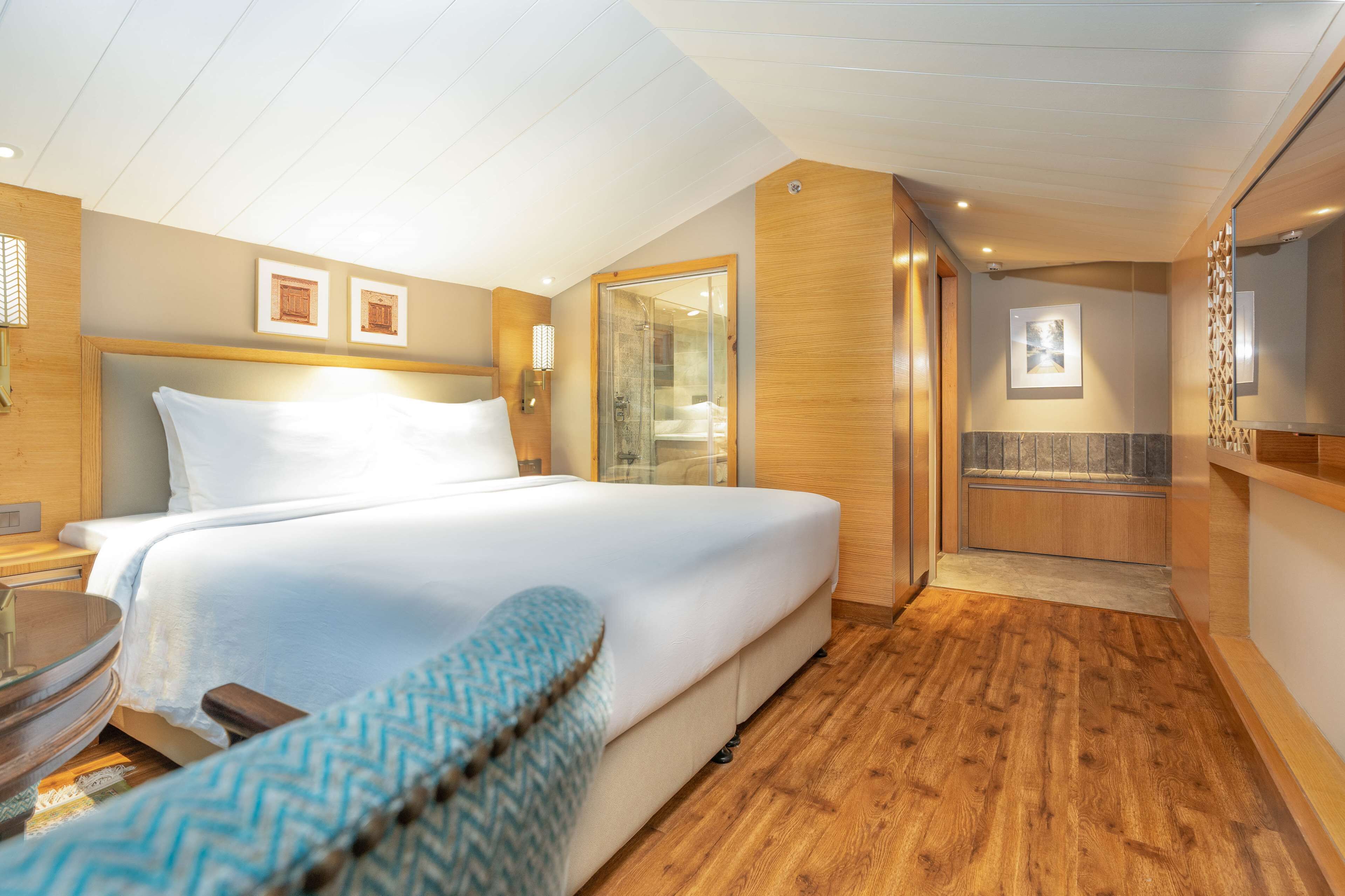 A hotel room with a large bed, a wooden floor, a glass shower enclosure, and a small seating area.