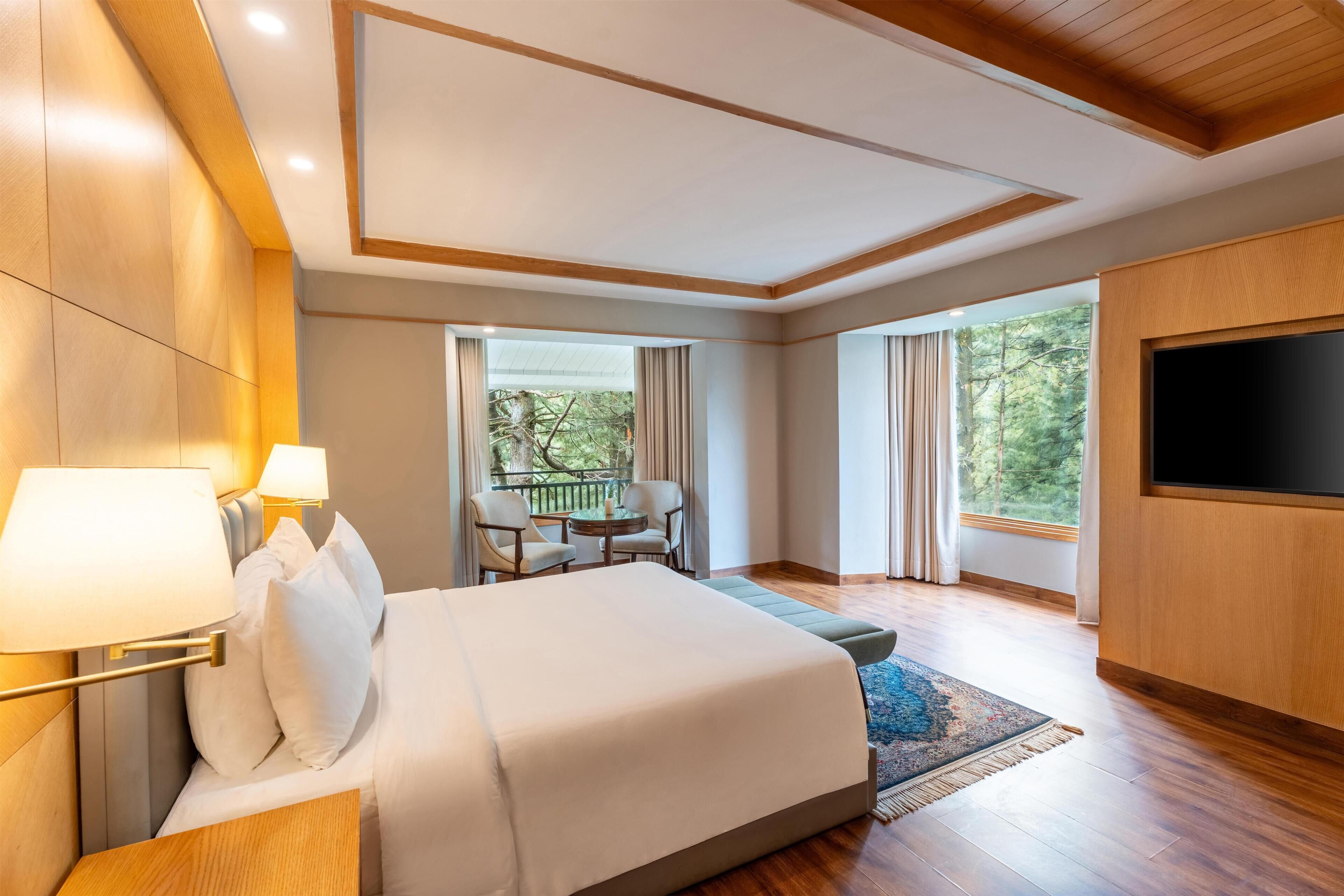 A hotel room with a bed, bedside lamps, a TV, a balcony with a table and chairs, and large windows.